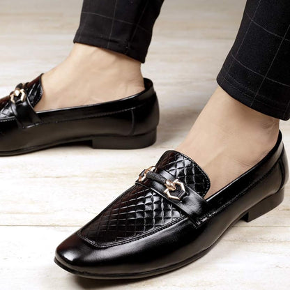 Black loafers with gold accents worn by a person on a light wooden floor.