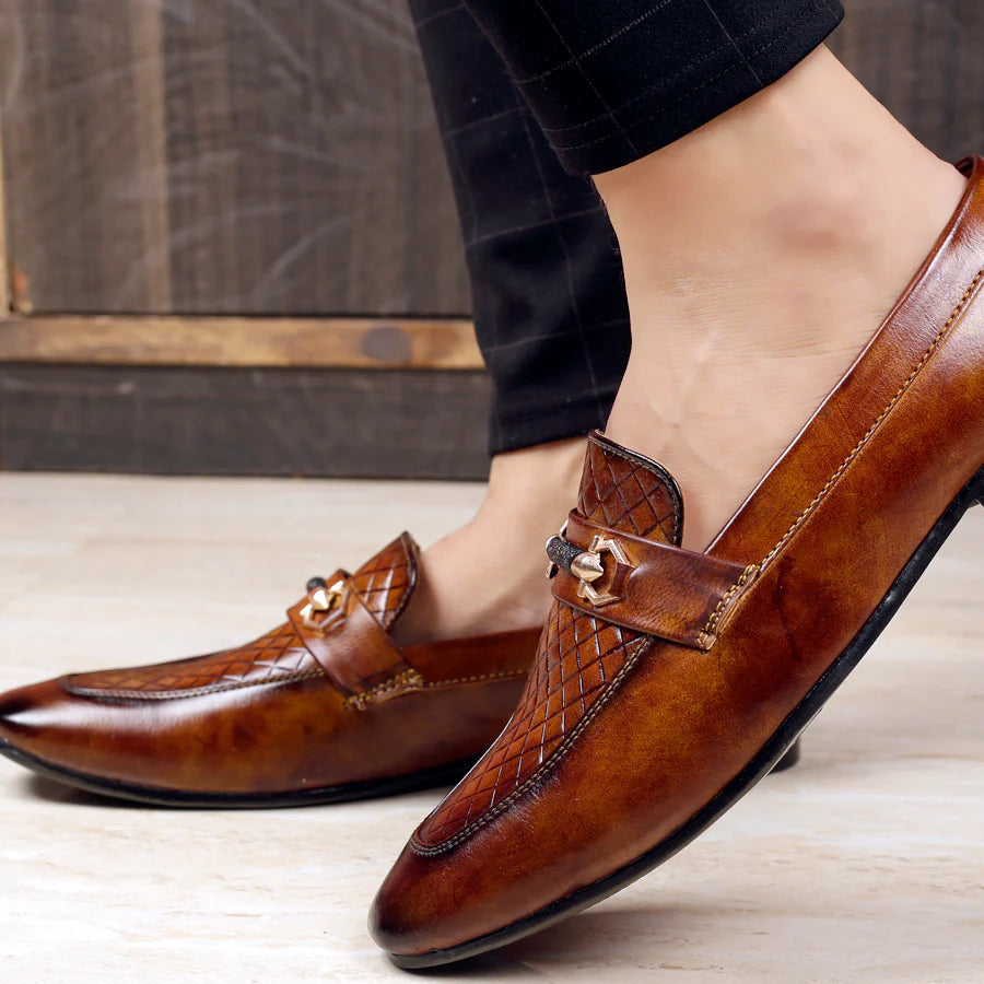 Brown leather loafers with decorative buckles worn by a person on a wooden floor.