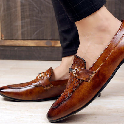 Brown leather loafers with decorative buckles worn by a person on a wooden floor.